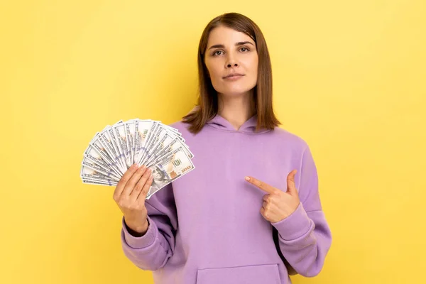 大金を持ってる 金持ちの若い大人の女性の肖像は ドル紙幣を指して 穏やかな自信を持って表現 財政の節約を探しています 黄色の背景に隔離された屋内スタジオショット — ストック写真