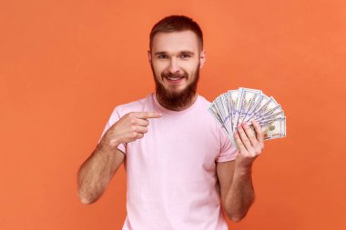 Dolar banknotlarını işaret eden sakallı bir adamın portresi ve mutlu bir ifadeyle, finansal tasarruflarla pembe tişört giyen bir adam. Turuncu arka planda kapalı stüdyo çekimleri.