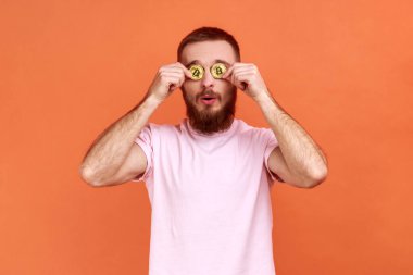 Şaşırmış sakallı adamın portresi altın bitcoin ile gözleri kaplayan, btc bozuk parayla kameraya bakan, pembe tişört giyen. Turuncu arka planda kapalı stüdyo çekimleri.