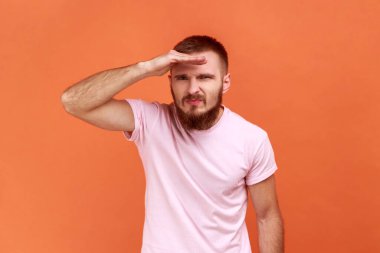 Uzaktan bakan sakallı bir adamın portresi başıyla dikkatle parlak bir gelecek arıyor, pembe tişört giyiyor. Turuncu arka planda kapalı stüdyo çekimleri.