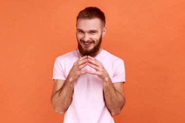 Kıskanç gülüşü olan, kurnaz, sakallı, yakışıklı bir adamın portresi entrikalar çeviren, hilekar numaralar yapan, pembe tişört giyen. Turuncu arka planda kapalı stüdyo çekimleri.