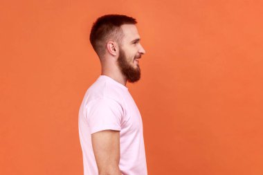 Sakallı adamın portresi ileriye bakıyor ve gülümsüyor, olumlu duygular ifade ediyor, iyi ruh hali içinde, pembe tişört giyiyor. Turuncu arka planda kapalı stüdyo çekimleri.