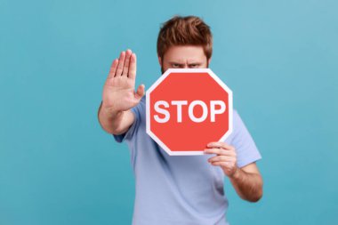 Yüzünün yarısını sekizgen dur sembolüyle kaplayan sakallı adamın portresi kırmızı trafik işareti ve yasak yol uyarısı ile el hareketini yasaklıyor. Mavi arka planda kapalı stüdyo çekimi.