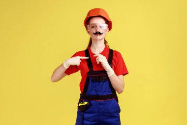 Kendine güvenen komik bir müteahhit kadının portresi çubuktaki bıyığı gösteriyor, kameraya bakıyor, tulum giyiyor ve koruyucu kask takıyor. Sarı arka planda kapalı stüdyo çekimleri.