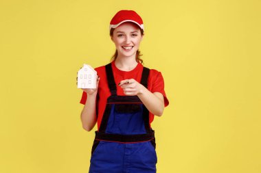 Elinde kağıt evle ayakta duran kadın nakliyecinin portresi ve kamera ev onarım ve onarım hizmetlerine işaret eden, tulumlu ve kırmızı şapkalı. Sarı arka planda kapalı stüdyo çekimleri.