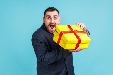 Sakallı, resmi stil takım elbiseli, hediye kutusu taşıyan heyecanlı bir adam, bayram hediyesiyle tatmin olmuş, hayaller gerçek oluyor, hoş sürprizler. Mavi arka planda kapalı stüdyo çekimi.