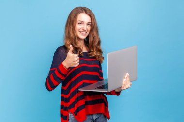 Güzel bir kadın, çizgili bir süveter giyiyor, elinde not defteri var ve baş parmağını kaldırıyor, serbest çalışan işlerinden memnun. Mavi arka planda kapalı stüdyo çekimi.