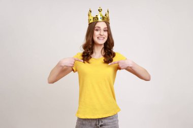 Sarı günlük tişörtlü ve kafasında altın taç olan neşeli genç kız liderlik vasıflarını gösteriyor dişlek bir gülümsemeyle kendini işaret ediyor. Gri arka planda izole edilmiş kapalı stüdyo görüntüleri.