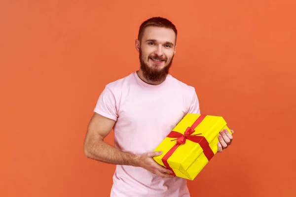 一个快乐的 积极的 留着胡子的男人站在那里 手里拿着黄色礼品盒 面带微笑地看着相机 穿着粉色T恤的画像 在橙色背景下被隔离的室内拍摄 — 图库照片