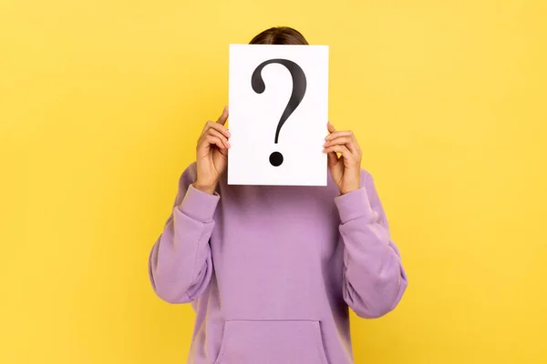 Portrait of shy woman hiding her face behind white paper with question mark, finding smart solution, asking for advice, wearing purple hoodie. Indoor studio shot isolated on yellow background.