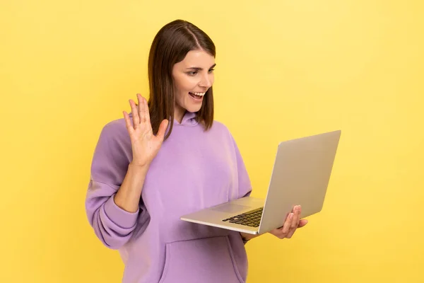 暗い髪を手を振って美しい女性は ノートパソコンの画面にこんにちはジェスチャー 友人とのビデオ通話 オンライン通信 紫色のパーカーを着ている 黄色の背景に隔離された屋内スタジオショット — ストック写真