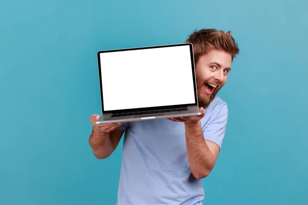白い画面でノートパソコンを持っている髭の男の肖像画と興奮顔の表情とおもちゃの笑顔でカメラを見て 青い背景に隔離された屋内スタジオショット — ストック写真