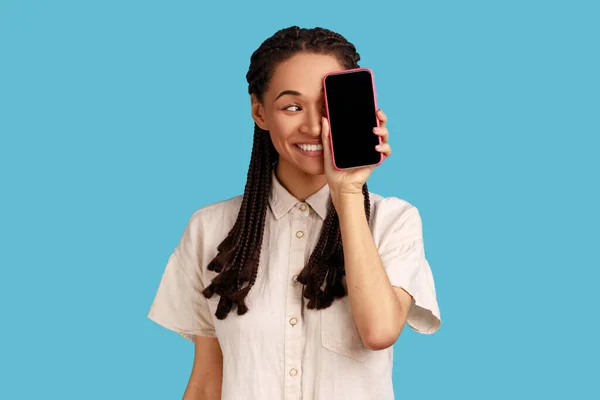 一个乐观的女人的画像 她用手机把半个人头盖住 用手机遮住眼睛 望着别处 表达着幸福 室内拍摄被蓝色背景隔离 — 图库照片