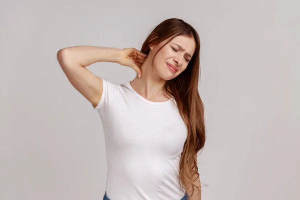 魅力的な疲れ動揺不健康な女性の肖像画背中の痛み 筋肉の緊張を緩和するためにマッサージ首立って 白いTシャツを着て 灰色の背景に隔離された屋内スタジオショット — ストック写真