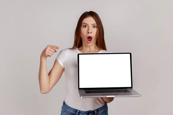 携帯コンピュータの白い空のディスプレイを指して暗い髪を持つショックを受けた女性 広告のためのコピースペース 白いTシャツを着て 灰色の背景に隔離された屋内スタジオショット — ストック写真