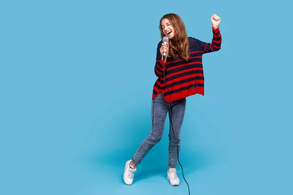 縞模様のカジュアルスタイルのセーターを着た女性の完全な長さの肖像画 マイクで歌い カラオケで楽しみを持っています 単独で実行 青い背景に隔離された屋内スタジオショット — ストック写真