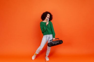 Afro saçlı, yeşil günlük süveter giyen, elinde teyp ve mikrofonla şarkı söyleyen bir kadının tam boy portresi. Turuncu arka planda kapalı stüdyo çekimleri.