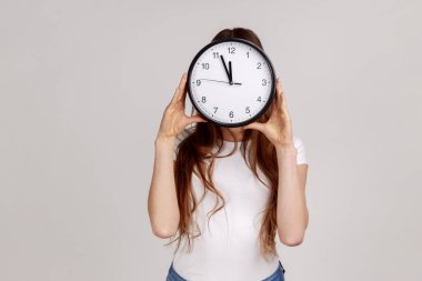 Bilinmeyen bir kadının duvar saatini tutan portresi, yüzünü gizleyen, zaman yönetimi, randevu ve beyaz tişört giyen. Gri arka planda izole edilmiş kapalı stüdyo görüntüleri.