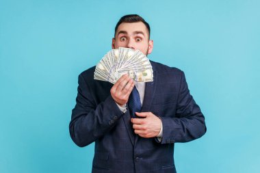 Yüzlük banknotların arkasına yüzünün yarısını saklayan koyu renk takım elbiseli bir işadamı büyük bir galibiyetle, bahisle şok oldu. Mavi arka planda kapalı stüdyo çekimi.