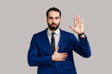 Katı sakallı bir adamın portresi bir kolunu kaldırıp göğsüne koyuyor diğeri yemin ediyor, küfrediyor, resmi tarz takım elbise giyiyor. Gri arka planda izole edilmiş kapalı stüdyo görüntüleri.