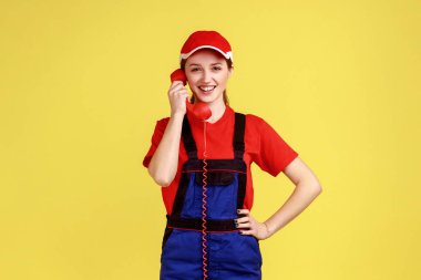 Müşterilerle kırmızı retro telefondan konuşan pozitif işçi kadın siparişleri kabul ediyor, profesyonel hizmet endüstrisi, tulum ve kırmızı şapka giyiyor. Sarı arka planda kapalı stüdyo çekimleri.