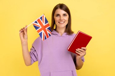 Çekici bir öğrenci portresi koyu renk saçlı kadın kitap tutuyor ve İngiltere bayrağı taşıyor, yurtdışında eğitim görüyor, mor kapüşonlu giyiyor. Sarı arka planda kapalı stüdyo çekimleri.