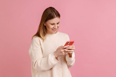 Akıllı telefon kullanan, cihaz ekranına bakan, yazı yazan, beyaz kazak giyen mutlu sarışın kadının portresi. Pembe arka planda kapalı stüdyo çekimleri.