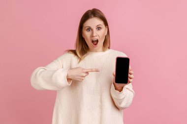 Cep telefonunu elinde tutan, terfi için fotokopi çekilen, beyaz süveter giyen akıllı telefon boş ekrana bakan hayretler içindeki bir kadının portresi. Pembe arka planda kapalı stüdyo çekimleri.