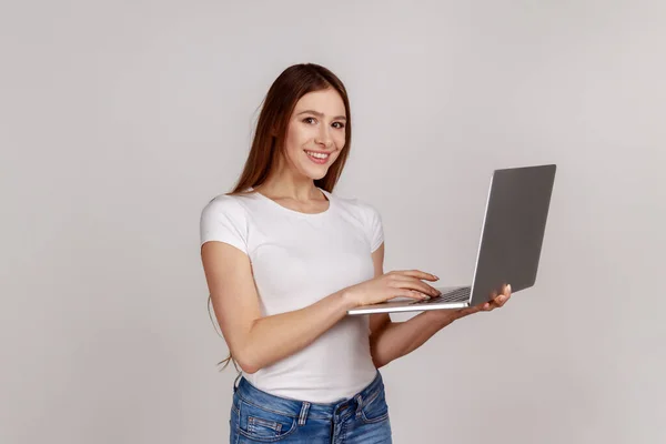 幸せな正の女性は カメラを見て ネットサーフィン 学生やフリーランスの仕事 コンピュータを使用して 白いTシャツを着て ラップトップを保持する 灰色の背景に隔離された屋内スタジオショット — ストック写真