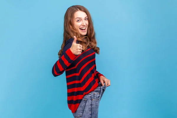 縞模様のカジュアルなスタイルのセーターを着て非常に幸せな女性の側面図は 親指を表示し 成功した減量を誇る 食事を維持します 青い背景に隔離された屋内スタジオショット — ストック写真