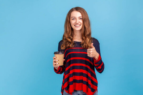 woman wearing striped casual style sweater, holding take away coffee and showing thumb up, recommend coffee house, excellent service.Indoor studio shot isolated on blue background.
