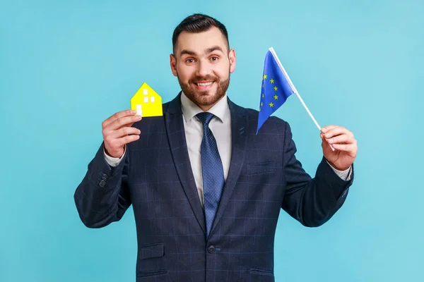 Good looking man with beard wearing official style suit holding model of paper house and europe union flag, dreaming about living in European country. Indoor studio shot isolated on blue background.
