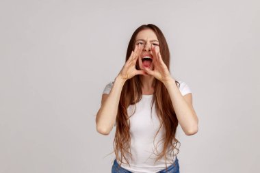 Siyah saçlı kızgın bir kadının portresi kötü haber diye bağırıyor ya da elleri ağzında beyaz tişört giyiyordu. Gri arka planda izole edilmiş kapalı stüdyo görüntüleri.