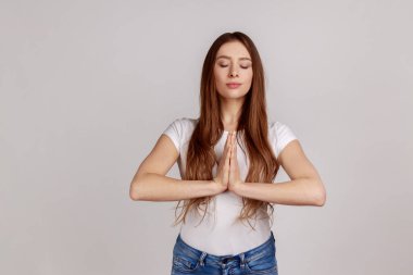 Sakin bir kadının portresi el ele tutuşması namaste jesti, meditasyon, yoga nefes alma tekniği, stresi azaltma, beyaz tişört giyme. Gri arka planda izole edilmiş kapalı stüdyo görüntüleri.