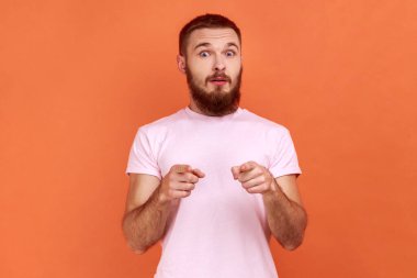 Şok olmuş sakallı adamın portresi büyük gözlü, ağzı açık, kamerayı işaret eden, senden şüphelenen, pembe tişört giyen. Turuncu arka planda kapalı stüdyo çekimleri.