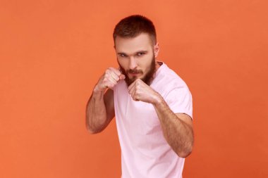 Sakallı agresif dövüşçünün portresi yumruklarını sıkmış boksa hazır, dövüş sanatları eğitmeni, kendini savunan, pembe tişört giyen. Turuncu arka planda kapalı stüdyo çekimleri.