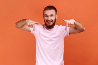 Keyifli tatmin olmuş, parmaklarını aşağı gösteren, reklam alanına dikkat eden, blogcu abone olmayı öneren, pembe tişört giyen bir adam. Turuncu arka planda kapalı stüdyo çekimleri.