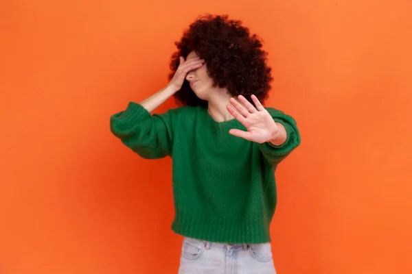 緑のカジュアルなスタイルのセーターを着て 手のひらで目をカバーし 手のひらで停止禁止ジェスチャーを示すアフロの髪型と落ち込んで女性 オレンジの背景に隔離された屋内スタジオショット — ストック写真