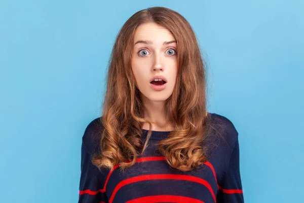 Wow, unbelievable! Surprised woman wearing striped casual style sweater has shocked expression, looking at camera with open mouth and amazed big eyes. Indoor studio shot isolated on blue background.