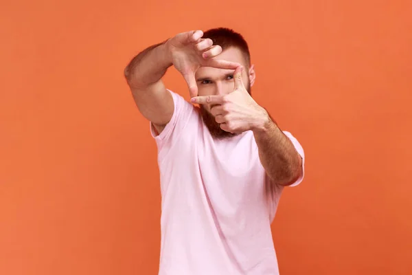 ピンクのTシャツを着て 適切なショット カメラマンの仕事を探して目に手を上げ フレームを作る集中的な創造的な髭の男の肖像画 オレンジの背景に隔離された屋内スタジオショット — ストック写真