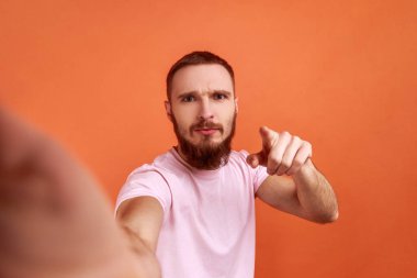 Ciddi derecede yakışıklı sakallı bir adamın selfie ya da akış yapan portresi, kameraya işaret eden, POV, fotoğrafın bakış açısı, pembe tişört giyen. Turuncu arka planda kapalı stüdyo çekimleri.