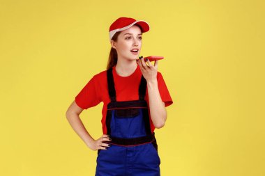Cep telefonunu elinde tutan ve müşteriye sesli mesaj kaydeden, tulumlu ve kırmızı şapkalı çekici bir kadının portresi. Sarı arka planda kapalı stüdyo çekimleri.