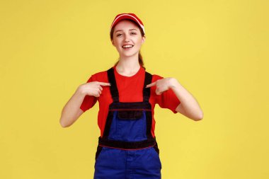 Ayakta duran ve kendini işaret eden mutlu işçi kadının portresi, işinden memnun olması, tulum giymesi ve kırmızı şapka takması. Sarı arka planda kapalı stüdyo çekimleri.