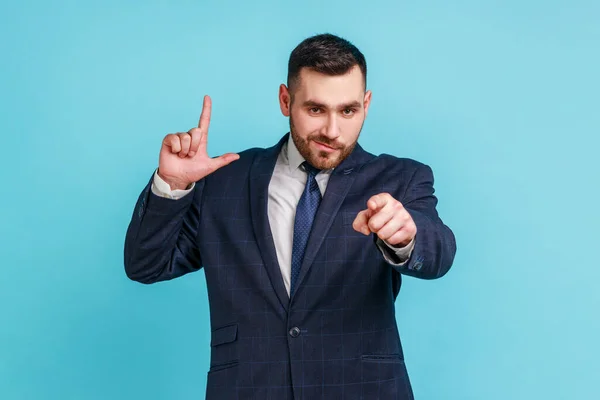 You lost! Portrait of dissatisfied businessman with beard in suit showing loser or lame gesture and pointing to camera, accusing for failure. Indoor studio shot isolated on blue background.