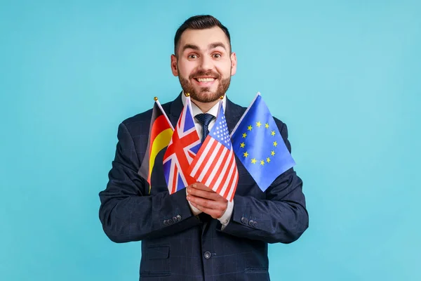 Positive man with beard wearing official style suit holding flags of different countries, language learning, looking at camera with toothy smile. Indoor studio shot isolated on blue background.