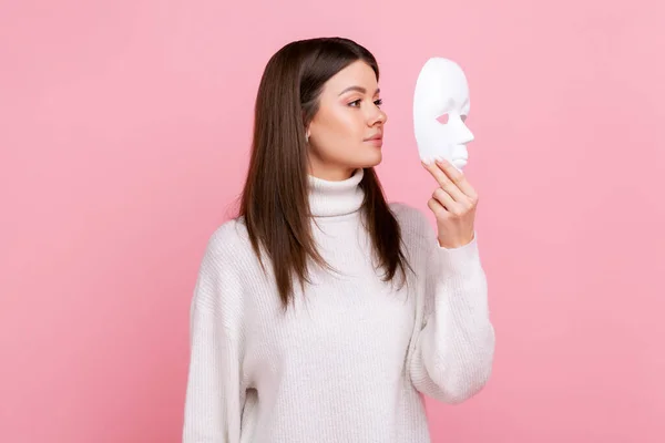 Side view portrait of girl holds white face mask, has mysterious expression, hiding his personality, wearing white casual style sweater. Indoor studio shot isolated on pink background.