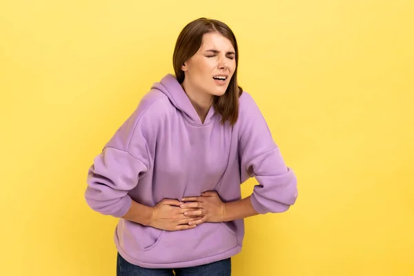 描述不健康的痛苦女人紧紧抓住她的腹部 由于急性腹痛 胃炎或便秘而面带笑容 穿着紫色的帽衫 在黄色背景下隔离的室内拍摄 — 图库照片