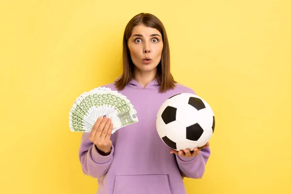 口を開いてサッカーボールを保持し ユーロのファンを持つ女性の肖像画 サッカー選手権の驚き 紫色のパーカーを着て 黄色の背景に隔離された屋内スタジオショット — ストック写真
