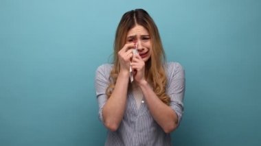 Kaşlarını çatan, gözyaşlarını silen, umutsuz hisseden, duygusal stresle baş eden, çizgili gömlek giyen sarışın bir kadının portresi. Mavi arka planda kapalı stüdyo çekimi.
