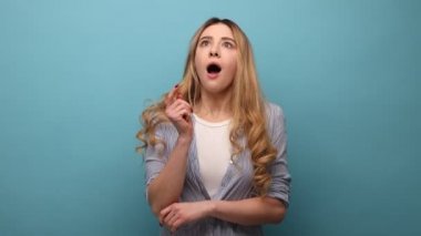 Aşırı neşeli, dalgalı sarı saçlı, parmağını kaldırıp kameraya bakan dişlek gülüşlü, fikri olan, iş planı olan, yeni başlayan, çizgili gömlek giyen bir kadın. Mavi arka planda kapalı stüdyo çekimi.